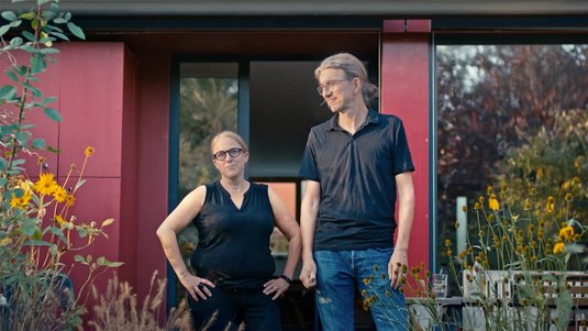 Vorschaubild für ENTEGA-Unternehmensfilm. Zwei Personen stehen vor einem modernen Gebäude mit roter Fassade, umgeben von Pflanzen.