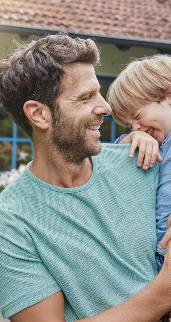 Lachender Vater trägt fröhlichen Jungen im Arm vor einem Haus mit blauem Fensterrahmen. Ausdruck von Freude und familiärer Verbindung an einem sonnigen Tag.