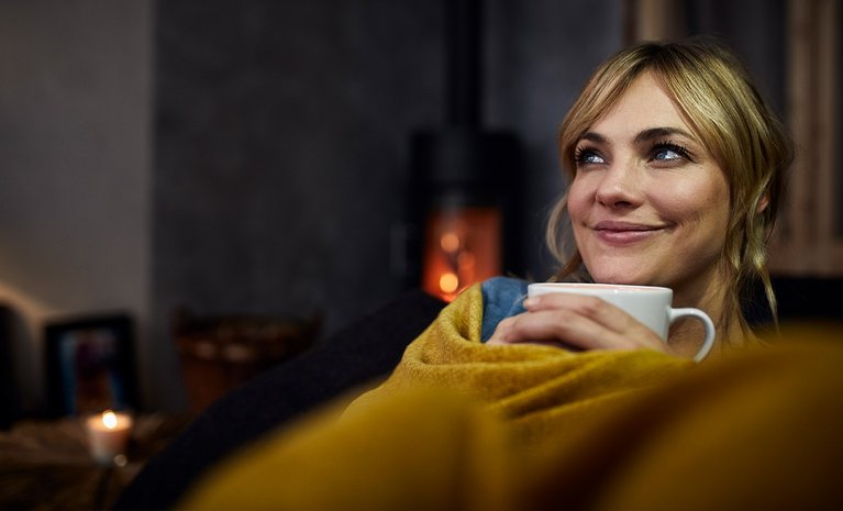 Frau sitzt lächelnd in einer gemütlichen Umgebung, eingehüllt in eine Decke, und hält eine Tasse. Ein Ofen brennt sanft im Hintergrund.