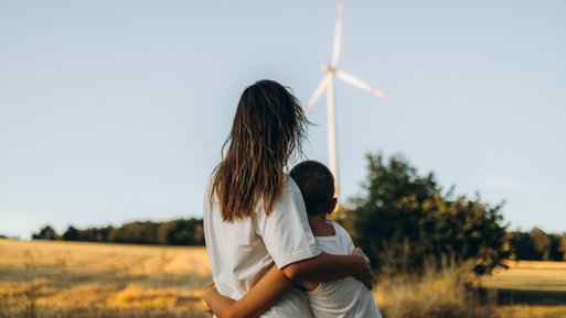 Zwei Personen umarmen sich auf einem Feld, im Hintergrund ein Windrad. Symbol für saubere Energie aus Windkraft.