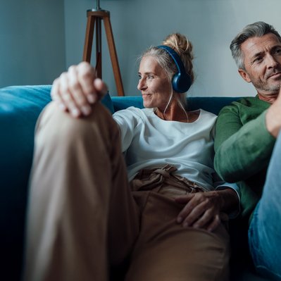Entspanntes Paar auf Sofa, Frau mit Kopfhörern, Mann mit Smartphone. Symbolisiert schnelles Internet und Telefonie mit ENTEGA Home Kombi.