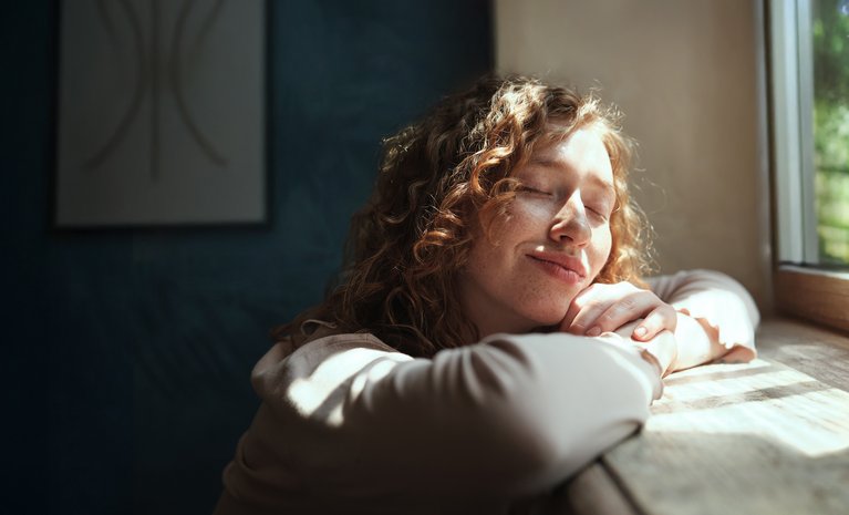 Frau, entspannt am Fenster im Sonnenlicht, strahlt Zufriedenheit aus. Vorteile einer gemieteten Wärmepumpe ohne hohe Anschaffungskosten.