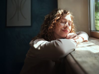 Frau, entspannt am Fenster im Sonnenlicht, strahlt Zufriedenheit aus. Vorteile einer gemieteten Wärmepumpe ohne hohe Anschaffungskosten.