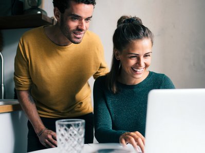 Fröhliches Paar schaut lachend auf einen Laptop in der Küche, eine Person in Gelb, die andere in Grün.