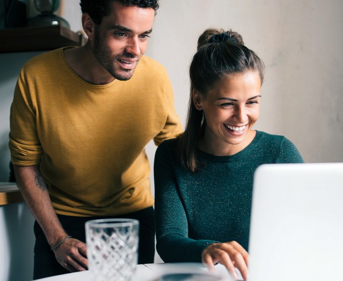 Fröhliches Paar schaut lachend auf einen Laptop in der Küche, eine Person in Gelb, die andere in Grün.