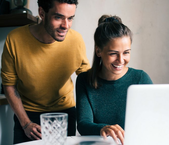 Fröhliches Paar schaut lachend auf einen Laptop in der Küche, eine Person in Gelb, die andere in Grün.