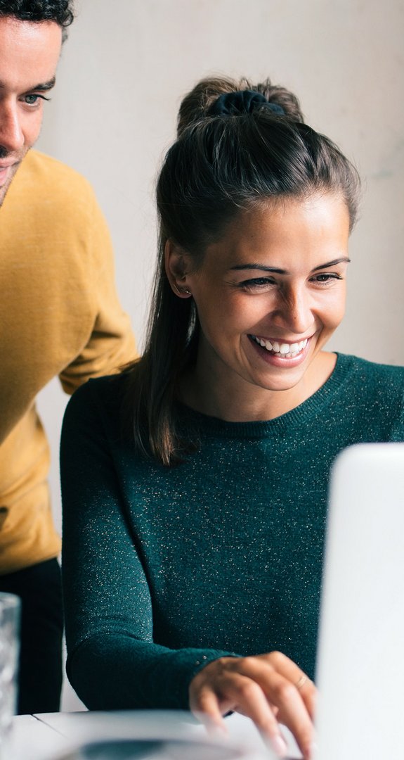 Fröhliches Paar schaut lachend auf einen Laptop in der Küche, eine Person in Gelb, die andere in Grün.
