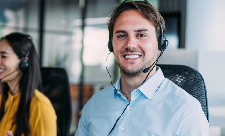 Ein lächelnder Mann mit Headset sitzt in einem offenen Büro, steht für professionelle und engagierte Unterstützung.