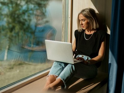 Frau sitzt an sonnigem Fenster mit Laptop, arbeitet konzentriert. Hohe Internetgeschwindigkeit durch Glasfaserausbau in Südhessen.