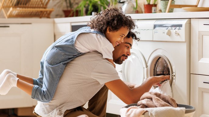 Bild zeigt Kind auf dem Rücken eines Mannes, der eine Waschmaschine befüllt – Thema: Stromverbrauch im Haushalt.