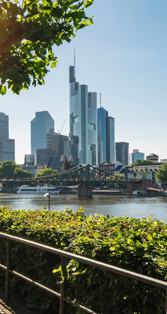 Frankfurts Skyline unter Sonnenschein, Fluss im Vordergrund. ENTEGA als klimafreundlicher Ökostromanbieter in Frankfurt am Main.