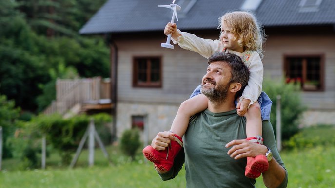 Lächelnder Mann mit Kind und Mini-Windrad vor Haus, steht für nachhaltigen ENTEGA Strommix mit weniger CO₂-Emissionen.
