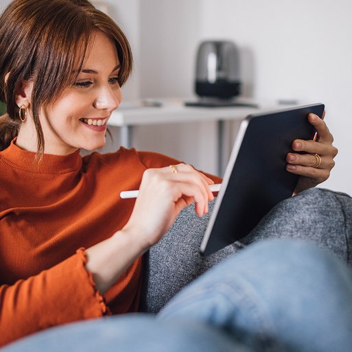 Frau sitzt entspannt auf einem Sofa und nutzt ein Tablet. Umgebung: modernes Wohnambiente mit Pflanzen. Zweck: Interesse an Wärmestrom-Tarif wecken.