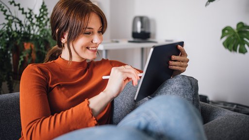 Frau sitzt entspannt auf einem Sofa und nutzt ein Tablet. Umgebung: modernes Wohnambiente mit Pflanzen. Zweck: Interesse an Wärmestrom-Tarif wecken.
