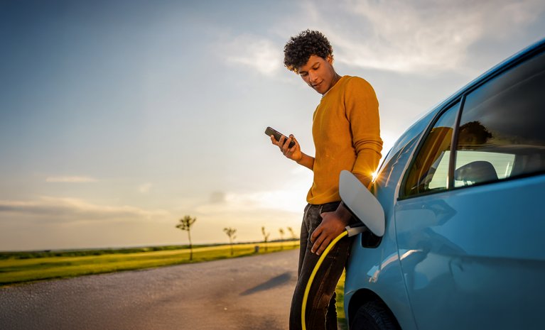 Person mit Handy neben Ladestation, lächelndes Gesicht bei Sonnenuntergang. Betont unkomplizierten Zugang zu vielen Ladepunkten.
