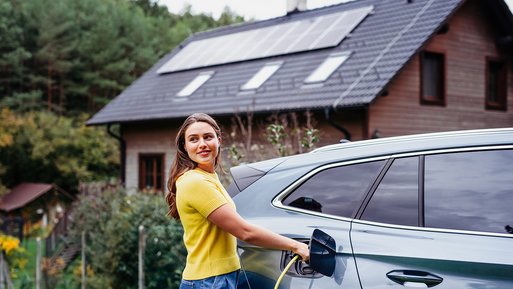 Frau lädt Elektroauto vor Haus mit Solarpanels. Eigenverbrauch von Solarstrom spart Kosten.