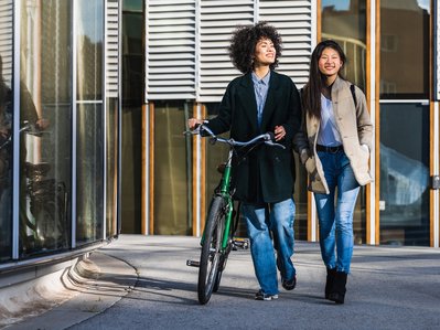 Zwei Frauen spazieren fröhlich an einem modernen Gebäude entlang, eine schiebt ein Fahrrad.