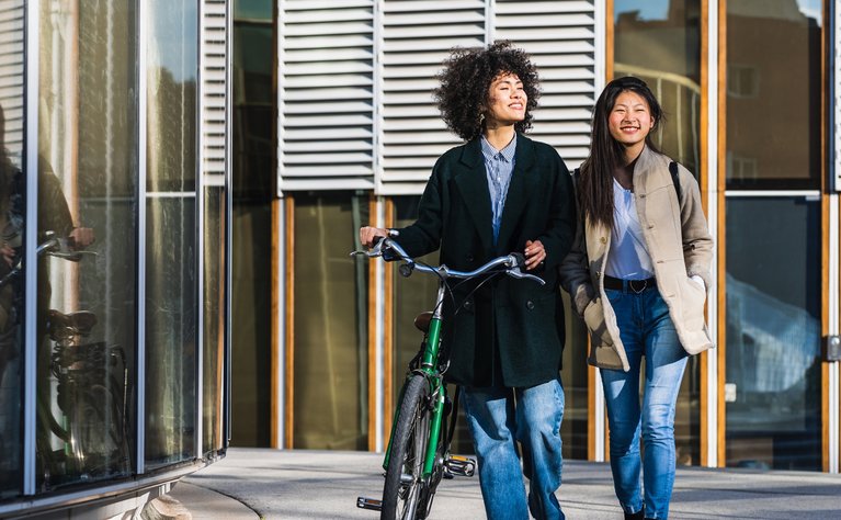 Zwei Frauen spazieren fröhlich an einem modernen Gebäude entlang, eine schiebt ein Fahrrad.