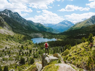 Nachhaltig reisen: Wanderer steht auf Felsblock vor Bergsee und Bergen, Fokus auf respektvollen Umgang mit Natur beim Reisen.
