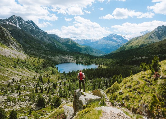 Nachhaltig reisen: Wanderer steht auf Felsblock vor Bergsee und Bergen, Fokus auf respektvollen Umgang mit Natur beim Reisen.