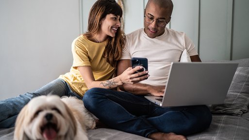 Paar sitzt mit Laptop und Smartphone auf dem Sofa, fröhlich; Hund liegt daneben. Kontext: Nutzung dynamischer Stromtarife zu Hause.