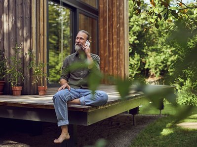 Mann sitzt gelassen auf einer Holzterrasse und telefoniert, umgeben von grünen Pflanzen. Symbolisiert Informationen über den einfachen Anbieterwechsel zu ENTEGA.