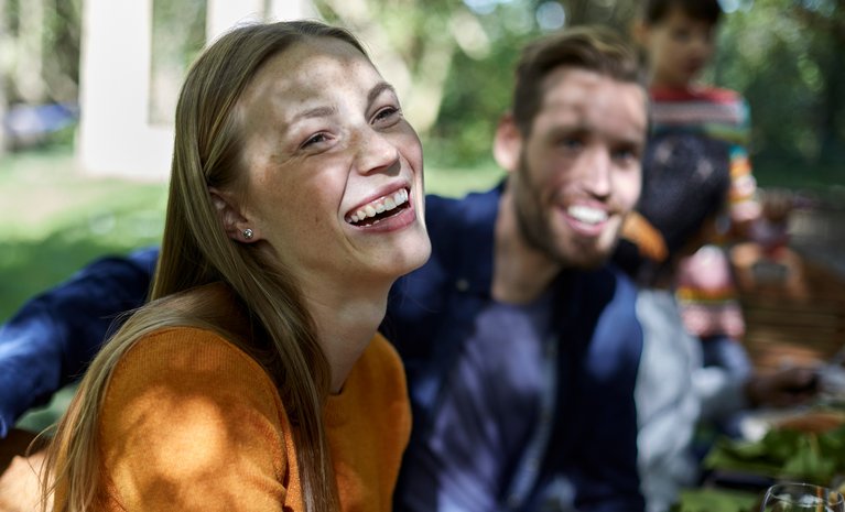 Lachende Frau draußen an einem Tisch mit Freunden, Sonnenlicht fällt durch Bäume. Ausdruck von Freude und Gemeinschaft im Freien.
