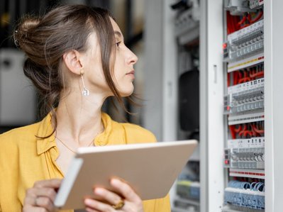 Frau mit Tablet inspiziert Elektroverteiler und die Funktionsweise des Stromzählers im Technikraum.
