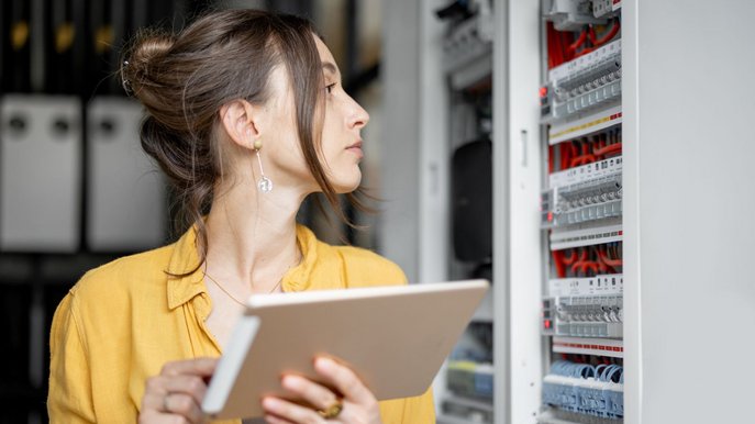 Frau mit Tablet inspiziert Elektroverteiler und die Funktionsweise des Stromzählers im Technikraum.