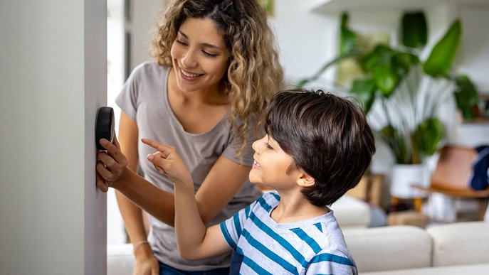 Frau und Junge stehen lachend vor einem digitalen Thermostat in einem Wohnzimmer. Sie justieren die Temperatur, um Energie zu sparen und Komfort zu erhöhen.