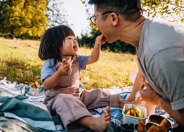 Kind füttert Erwachsene auf Picknickdecke mit Gemüse-Sticks; naturnahe Szene steht für nachhaltigen Lebensstil und Energiewende in Deutschland.