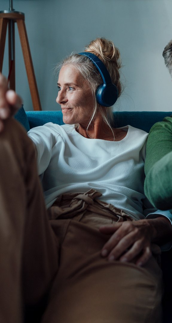 Entspanntes Paar auf Sofa, Frau mit Kopfhörern, Mann mit Smartphone. Symbolisiert schnelles Internet und Telefonie mit ENTEGA Home Kombi.