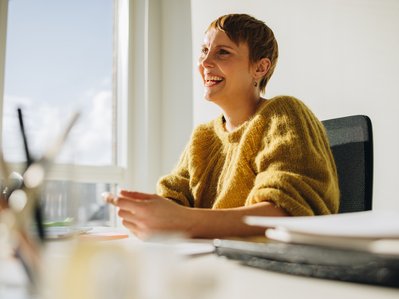 Eine Person in gelbem Pullover sitzt lächelnd an einem sonnigen Fenster im Büro, symbolisiert positive Geschäftsumgebung.