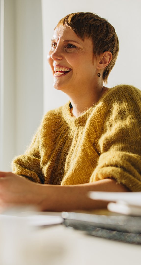 Eine Person in gelbem Pullover sitzt lächelnd an einem sonnigen Fenster im Büro, symbolisiert positive Geschäftsumgebung.