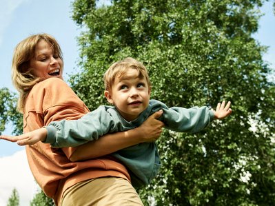 Kind fliegt spielerisch in den Armen einer Person vor grünem Hintergrund; Fortbewegungsmittel der Zukunft stehen für Freiheit und Visionen.