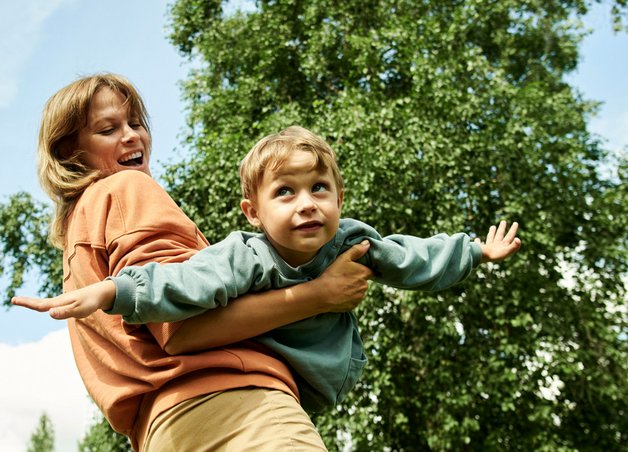 Kind fliegt spielerisch in den Armen einer Person vor grünem Hintergrund; Fortbewegungsmittel der Zukunft stehen für Freiheit und Visionen.