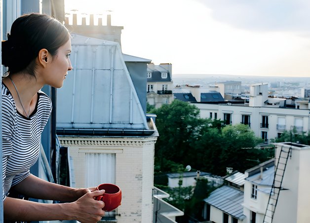 Bild zeigt Frau mit Kaffeetasse in der Hand, die aus dem Fenster auf eine Stadt blickt – Thema: klimafreundlich leben.