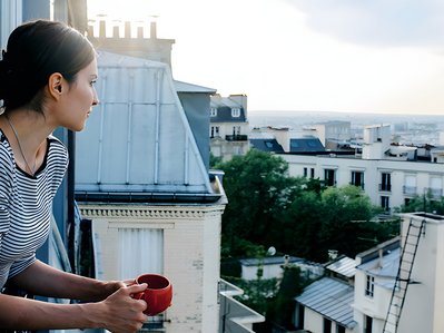 Bild zeigt Frau mit Kaffeetasse in der Hand, die aus dem Fenster auf eine Stadt blickt – Thema: klimafreundlich leben.
