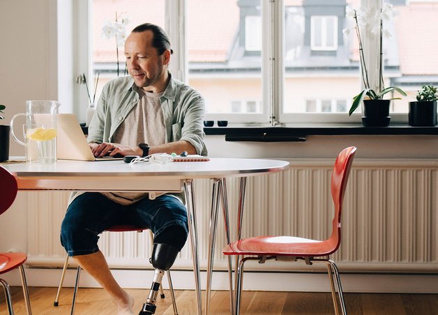 Mann mit Beinprothese arbeitet am Laptop in heller Küche, umgeben von Pflanzen. Thema: Was ist Heizstrom und wie kann er im Alltag genutzt werden?