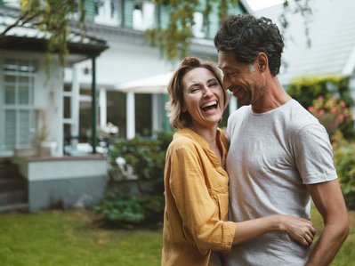 Zwei Menschen lächeln fröhlich und genießen die Zeit zusammen im Freien, im Hintergrund ein Wohnhaus.