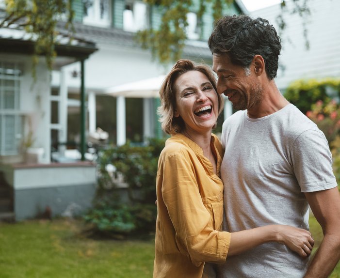 Zwei Menschen lächeln fröhlich und genießen die Zeit zusammen im Freien, im Hintergrund ein Wohnhaus.