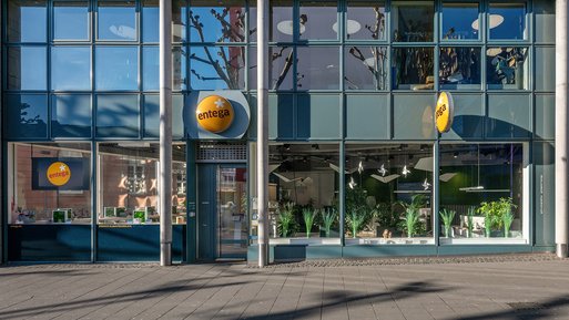 Gebäudeansicht des ENTEGA Servicepoints mit Glasfassade, gelbem Firmenlogo und sichtbaren Pflanzen im Inneren. Die Eingangstür befindet sich links.