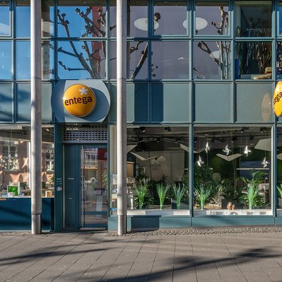 Gebäudeansicht des ENTEGA Servicepoints mit Glasfassade, gelbem Firmenlogo und sichtbaren Pflanzen im Inneren. Die Eingangstür befindet sich links.