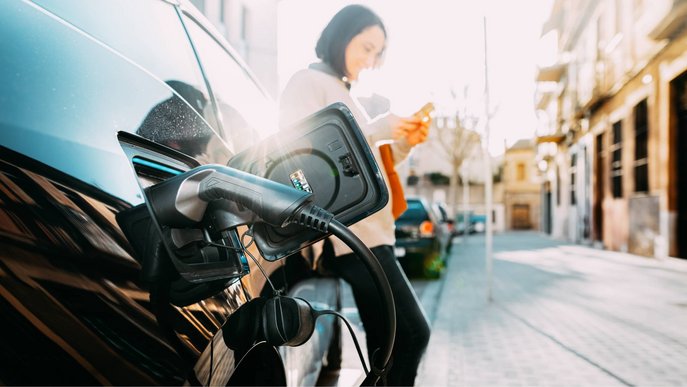 Bild zeigt Frau, die ihr Elektroauto lädt und ihr Handy nutzt – Thema: Tipps und Wissenswertes zur Elektromobilität.