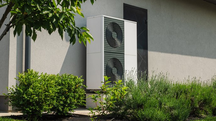 Wärmepumpe an der Fassade eines Hauses, umgeben von grünen Büschen. Symbolisiert umweltfreundliche und energieeffiziente Heiztechnologie.