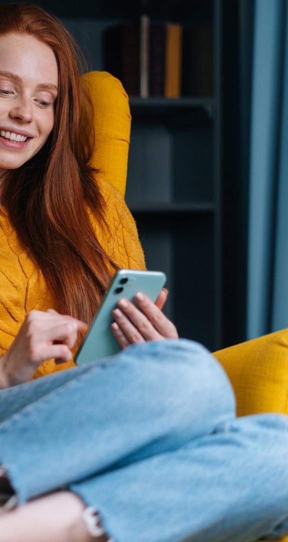 Frau in gelbem Pullover sitzt lächelnd auf einem gelben Sessel und nutzt ein Smartphone. Blaues Regal und Vorhang im Hintergrund.