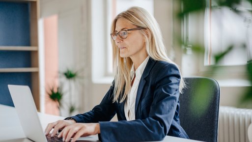 Eine Frau sitzt lächelnd am Fenster in einem modernen Büro, ihr Laptop vor sich, schafft eine produktive und angenehme Atmosphäre.
