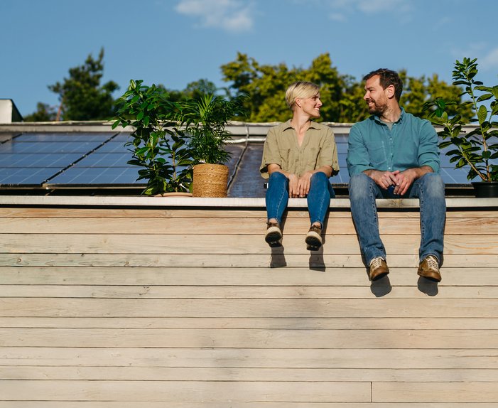 Paar sitzt lächelnd vor Solarmodulen auf dem Dach, umgeben von Pflanzen. Darstellung von Unabhängigkeit und Selbstversorgung mit Solarenergie.