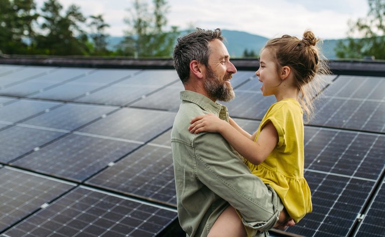 Ein Mann hält ein fröhliches Kind in gelbem Kleid vor einer großen Solaranlage. Sie lachen sich an, umgeben von Bäumen und Bergen.