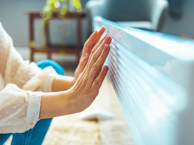 Bild zeigt eine Frau, die ihre Hände an einer Heizung wärmt – Elektroheizungen bieten vor allem in der Übergangszeit schnell verfügbare Wärme.